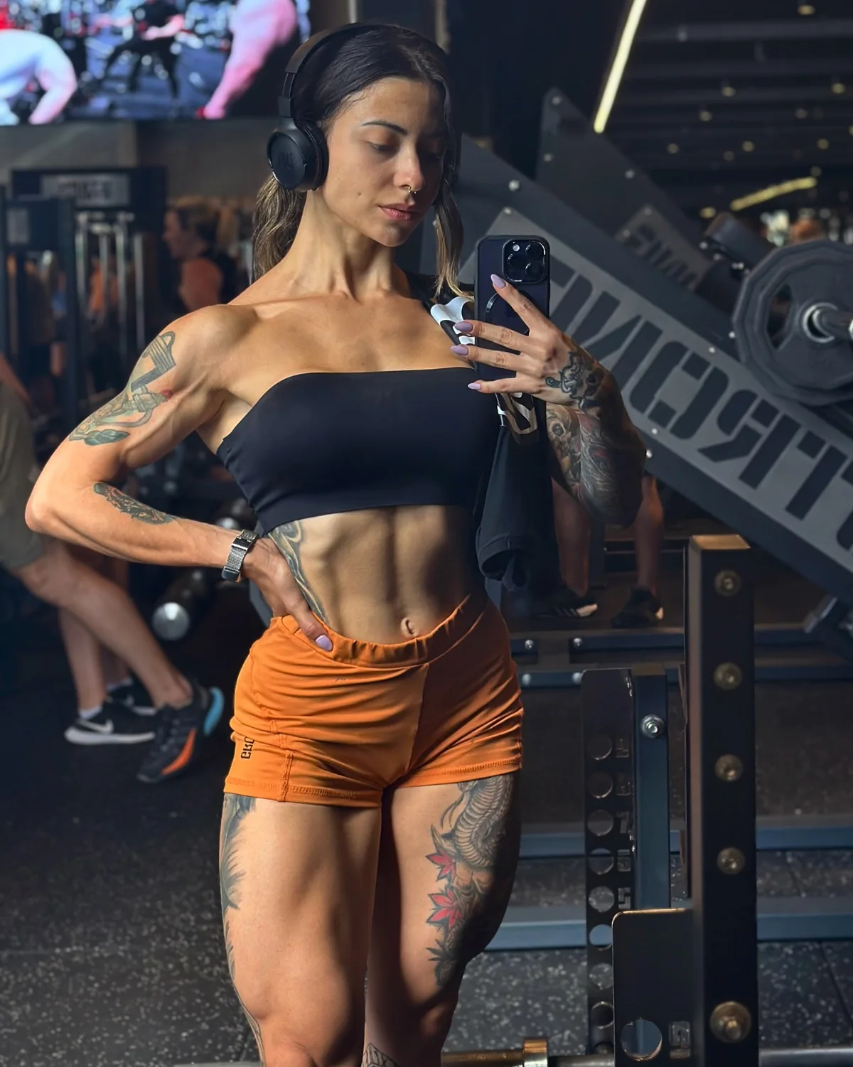 Bianca Mattos em selfie de espelho, de top preto e shorts laranja, na academia