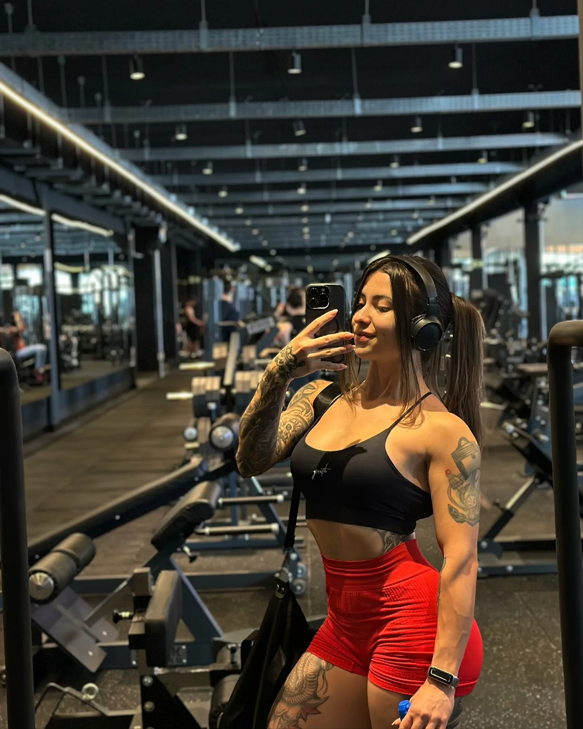 Bianca Mattos em selfie de espelho em academia grande, de top preto e shorts vermelhos