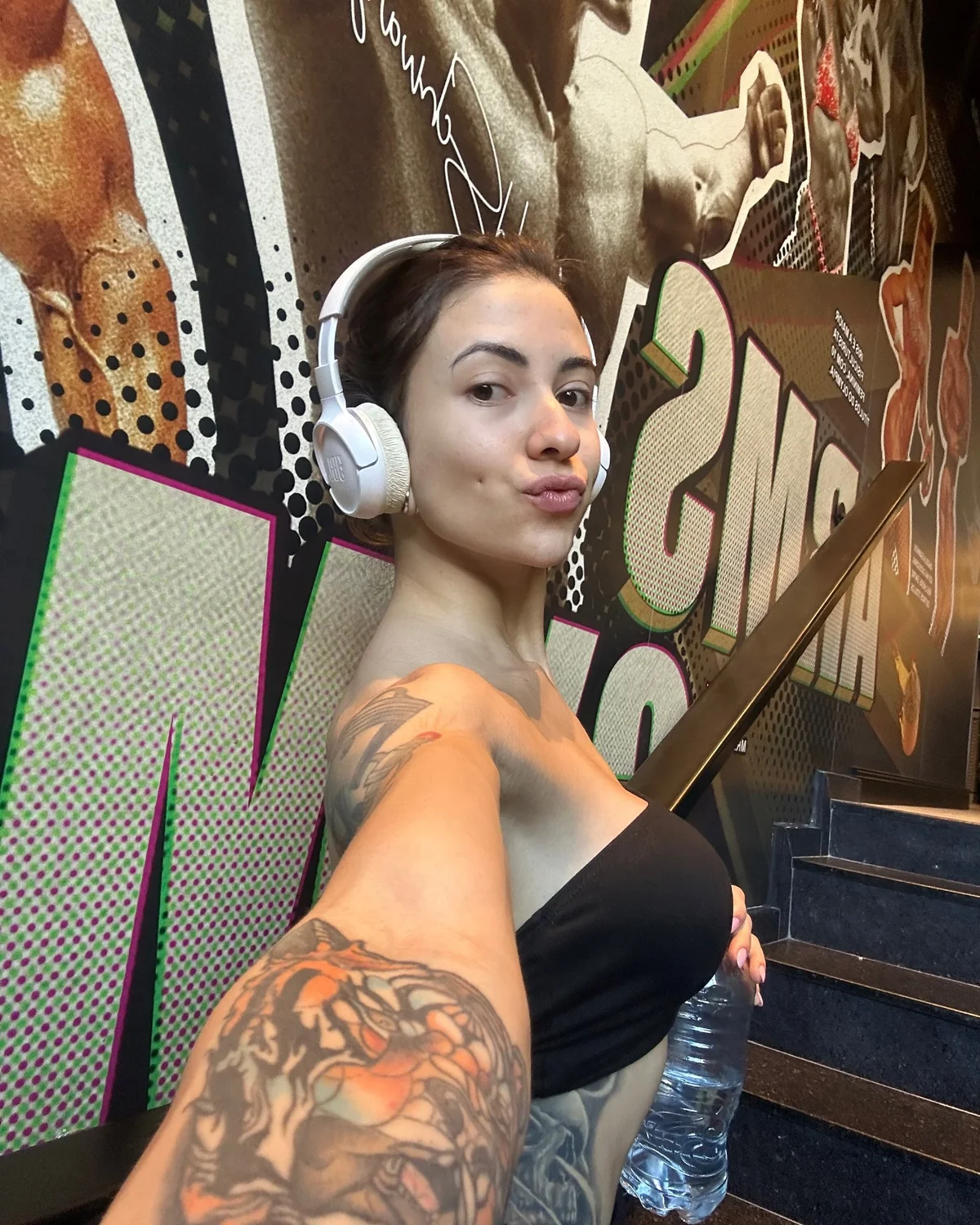 Bianca Mattos em selfie diante de um mural de academia, com fones brancos